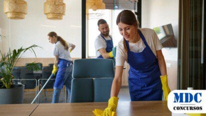 Três funcionários da limpeza, usando aventais azuis e luvas amarelas, higienizam um escritório moderno. Uma mulher ao centro limpa uma mesa de madeira, enquanto outro funcionário passa um pano em um vidro e uma mulher ao fundo aspira o chão. O ambiente é iluminado e decorado com plantas e luminárias / Cafelândia (PR) oferece 23 Vagas em Processo Seletivo
