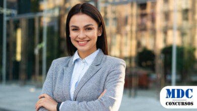 Mulher jovem de cabelos escuros e lisos sorri com os braços cruzados, vestindo um blazer cinza e camisa social listrada. Ela está em um ambiente urbano, com um prédio de vidro ao fundo refletindo a cidade - Câmara Municipal de Pratânia (SP) abre concurso com remuneração de até R$ 6,4 mil