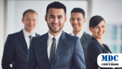Grupo de quatro profissionais em trajes formais, sorrindo em um ambiente corporativo. O homem na frente, de terno azul-marinho e gravata preta, é o foco da imagem. Ao fundo, levemente desfocados, estão dois homens e uma mulher, todos também bem vestidos e com expressões amigáveis - Câmara de Uberaba Oferece Remuneração de até R$ 5,2 mil em Concurso