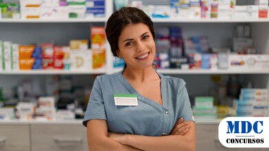 Processos Seletivos 9 Mulher de avental cinza, sorridente, de braços cruzados, em uma farmácia. Ela usa um crachá sem identificação. Ao fundo, prateleiras organizadas com caixas de medicamentos e produtos farmacêuticos. O ambiente é bem iluminado, e a profissional transmite simpatia e confiança - Campo Bom (RS) Lança PSS para Estagiários de Farmácia Bolsa Auxílio de até R$ 1,7 mil