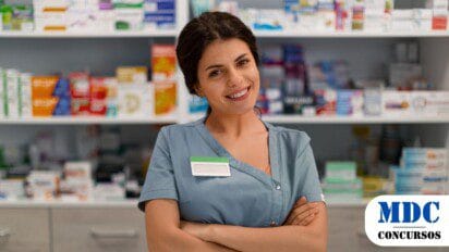 Mulher de avental cinza, sorridente, de braços cruzados, em uma farmácia. Ela usa um crachá sem identificação. Ao fundo, prateleiras organizadas com caixas de medicamentos e produtos farmacêuticos. O ambiente é bem iluminado, e a profissional transmite simpatia e confiança - Campo Bom (RS) Lança PSS para Estagiários de Farmácia Bolsa Auxílio de até R$ 1,7 mil