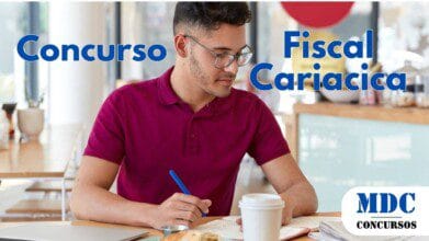 Homem jovem de óculos e camisa polo bordô estudando em um café, escrevendo em um caderno com caneta azul. Sobre a mesa, há um laptop, café em copo descartável, croissant em um prato e jornal. O ambiente é iluminado, com decoração moderna e janelas amplas ao fundo - Cariacica (ES) Abre 15 Vagas com Salário de R$ 3,3 Mil em Concurso