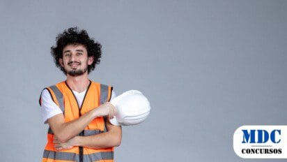 Homem jovem com colete de segurança laranja e capacete branco na mão, posando sorridente contra um fundo cinza neutro / Carneirinho - MG abre 30 vagas com salários de até R$ 6,3 mil em novo Processo Seletivo