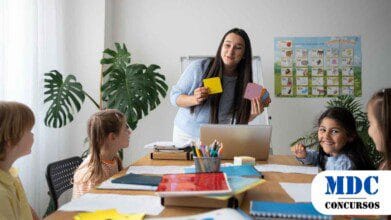 Professora segurando cartões coloridos enquanto ensina um grupo de crianças em uma sala de aula com materiais escolares sobre a mesa, um laptop e um pôster do alfabeto na parede/Chapadão do Céu (GO) Lança Processo Seletivo com 36 Vagas na Educação