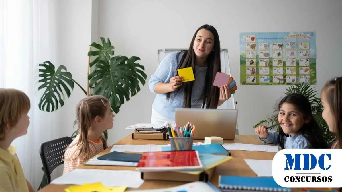Professora segurando cartões coloridos enquanto ensina um grupo de crianças em uma sala de aula com materiais escolares sobre a mesa, um laptop e um pôster do alfabeto na parede/Chapadão do Céu (GO) Lança Processo Seletivo com 36 Vagas na Educação