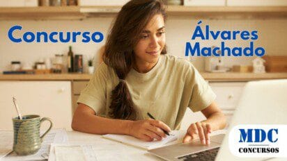 Mulher jovem com cabelo castanho preso de lado estuda em uma cozinha bem iluminada, escrevendo em um caderno enquanto usa um laptop, com uma xícara e documentos na mesa - Concurso Álvares Machado (SP) Tem Salários de R$ 5 mil