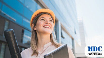 Mulher sorridente com capacete de segurança segurando uma pasta e um tubo de projetos em frente a um prédio moderno de vidro. A imagem transmite profissionalismo e confiança, sugerindo uma engenheira ou arquiteta no ambiente de trabalho / Concurso CAU/MG oferece 6 Vagas e Salários de até R$ 9,7 Mil
