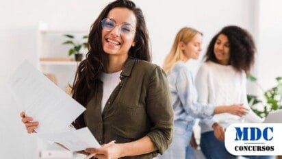 Mulher sorridente de óculos e cabelos longos segura documentos em um ambiente de escritório moderno, com duas colegas ao fundo conversando e usando um laptop - Concurso Câmara de Morrinhos GO 19 Vagas e Salários de até R$ 5,1 Mil