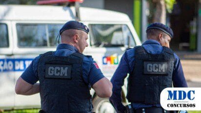 Dois guardas civis municipais (GCM) de uniforme azul escuro e coletes à prova de balas patrulham uma área urbana durante o dia, com um veículo branco ao fundo. Eles usam boinas e equipamentos táticos, incluindo rádios e coldres, caminhando lado a lado em uma rua movimentada - Concurso Coelho Neto (MA) Abre 120 Vagas para Guarda Municipal