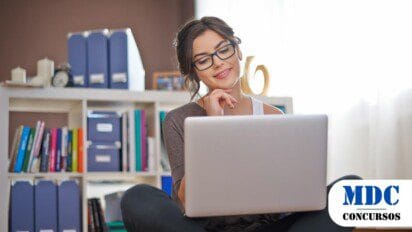 Mulher jovem de óculos sentada no chão, usando um laptop e sorrindo. Ao fundo, uma estante branca com livros, pastas e caixas organizadoras. O ambiente é iluminado por luz natural vinda de uma janela. Ela parece concentrada e confortável, transmitindo um clima de produtividade e bem-estar - Concurso Iaciara (GO) abre 171 Vagas em Diversas Áreas de Atuação