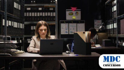 Mulher de terno bege trabalha em um laptop em um arquivo corporativo cercado por prateleiras com pastas organizadas, enquanto um colega ao fundo escreve em um caderno - Concurso PC CE 2025 100 Vagas para Delegado com Remuneração de R$ 22.165,53