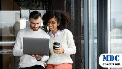 Homem e mulher sentados próximos a uma janela, sorrindo enquanto olham para um laptop. Ele veste um suéter branco, e ela usa óculos, suéter bege e calça rosa, segurando um café. O ambiente é moderno, com paredes de vidro e iluminação natural / Concurso Prefeitura de Tietê (SP) tem Salário de R$ 5,7 mil