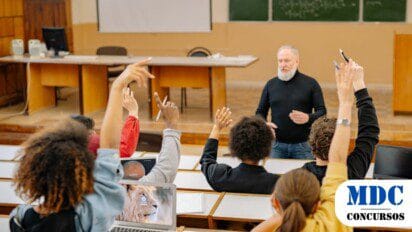 Professor de barba branca, vestindo suéter preto e jeans, dá aula em uma sala universitária. Estudantes levantam as mãos para fazer perguntas. Alguns usam laptops, enquanto outros seguram canetas. O ambiente tem um quadro negro com anotações matemáticas e uma mesa de professor ao fundo. A sala possui um estilo clássico de auditório / Concurso UFDPar abre vaga para Docente com remuneração de até R$ 13,3 mil