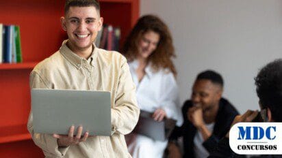 Jovem sorridente com camisa bege segura um laptop em um ambiente moderno com estante vermelha cheia de livros. Ao fundo, três pessoas interagem, uma delas tirando foto com o celular / Concurso UFSB e UFOB