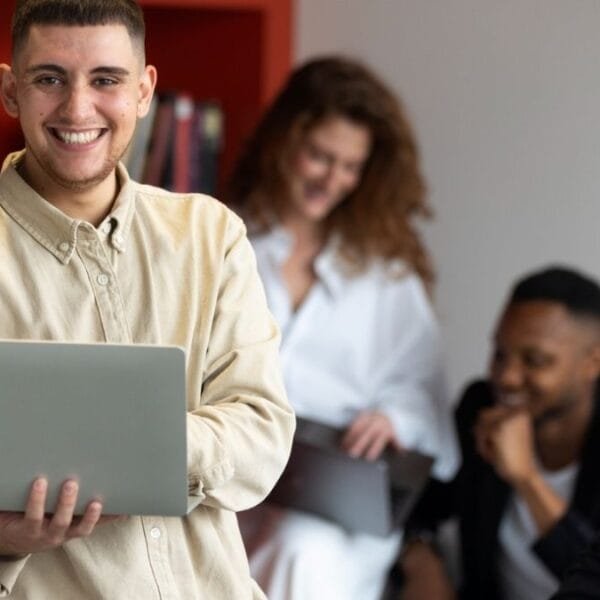 Jovem sorridente com camisa bege segura um laptop em um ambiente moderno com estante vermelha cheia de livros. Ao fundo, três pessoas interagem, uma delas tirando foto com o celular / Concurso UFSB e UFOB