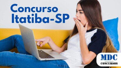 Mulher jovem de cabelos longos e castanhos sentada em um sofá amarelo, usando jeans e camiseta branca com preto, segurando um laptop no colo. Ela está boquiaberta, com uma mão no rosto e a outra apontando para a tela, expressando surpresa. Há almofadas azul e preta atrás dela - Concurso em Itatiba (SP) Oferece 286 Vagas com Salários de até R$ 10,8 Mil