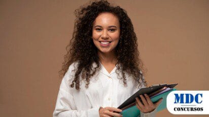 Mulher jovem de cabelos cacheados e longos sorrindo, vestindo camisa branca e calça preta, segurando uma prancheta e pastas coloridas em um fundo bege - Defensoria Pública do Amapá abre 20 vagas para Estágio em Direito