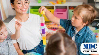 Professora sorridente com óculos e camiseta branca interage com crianças em uma sala de aula colorida. Uma criança loira de camisa jeans empilha blocos de madeira enquanto outras observam e participam. Ao fundo, caixas organizadoras e materiais educativos criam um ambiente lúdico e acolhedor - Educação de Canto do Buriti (PI) lança PSS com 48 Vagas + CR