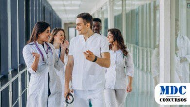 Processos Seletivos 6 Grupo de jovens profissionais da saúde caminhando por um corredor de hospital. Eles vestem jalecos brancos e roupas médicas, com estetoscópios no pescoço. Estão sorrindo e conversando de forma animada, transmitindo um ambiente positivo e descontraído. O corredor tem paredes de vidro refletindo a equipe / GDF abre 250 vagas com remuneração de até R$ 10 mil em PSS para Saúde no Distrito Federal