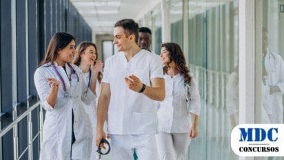 Grupo de jovens profissionais da saúde caminhando por um corredor de hospital. Eles vestem jalecos brancos e roupas médicas, com estetoscópios no pescoço. Estão sorrindo e conversando de forma animada, transmitindo um ambiente positivo e descontraído. O corredor tem paredes de vidro refletindo a equipe / GDF abre 250 vagas com remuneração de até R$ 10 mil em PSS para Saúde no Distrito Federal