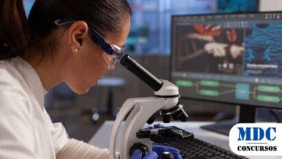 Cientista mulher de jaleco branco, óculos de proteção e luvas azuis observa uma amostra em um microscópio em um laboratório. Ao fundo, um monitor exibe imagens ampliadas e fluxogramas científicos. O ambiente é moderno, com equipamentos avançados e iluminação adequada para pesquisa - IDTECH abre Processo Seletivo para Rede HEMO em Goiás com salários de até R$ 5 mil