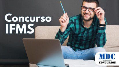 Homem sorridente de barba e óculos segurando um lápis enquanto ajusta os óculos, usando camisa xadrez verde e jeans, sentado em um sofá bege com um notebook e papéis sobre uma mesa azul clara, fundo escuro - IFMS oferece 78 Vagas e Remuneração de até R$ 13,3 Mil em novo Concurso
