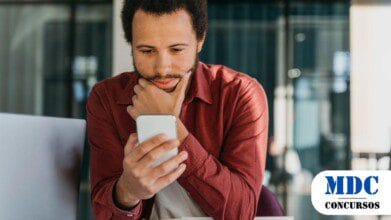 Homem jovem de barba curta e cabelos cacheados olha atentamente para o celular com expressão pensativa, segurando o queixo com a mão. Ele veste uma camisa vermelha e está sentado em um ambiente de escritório moderno com um laptop ao fundo - Ipeúna (SP) lança Concurso com 6 Vagas e Salários de até R$ 1.615,43