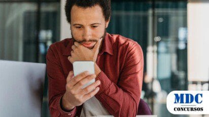 Homem jovem de barba curta e cabelos cacheados olha atentamente para o celular com expressão pensativa, segurando o queixo com a mão. Ele veste uma camisa vermelha e está sentado em um ambiente de escritório moderno com um laptop ao fundo - Ipeúna (SP) lança Concurso com 6 Vagas e Salários de até R$ 1.615,43