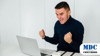 Homem jovem de suéter preto sentado à mesa, comemorando animado em frente a um laptop com uma xícara de café ao lado - Iporã do Oeste (SC) oferece Salários de até R$ 4,7 mil em Processo Seletivo