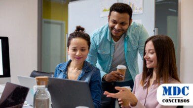 Três colegas de trabalho estão em um escritório colaborando em um projeto. Duas mulheres estão sentadas em frente a laptops, enquanto um homem de camisa azul está de pé, segurando um café e sorrindo. Eles parecem engajados e entusiasmados. O ambiente tem uma mesa com copos de café, garrafa de água e anotações / Itupeva (SP) Abre Concurso para Diversos Cargos