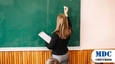 Machadinho D'Oeste (RO) Publica Resultado Final de Processo Seletivo com 247 Aprovados - Professora de costas escrevendo no quadro verde com giz branco, segurando uma prancheta na outra mão, enquanto duas crianças sentadas observam. A sala tem parede clara e lambril de madeira