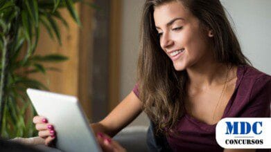 Mulher jovem com cabelos castanhos longos e ondulados, sorrindo enquanto usa um tablet. Ela veste uma blusa roxa e está sentada confortavelmente em um sofá. Suas unhas estão pintadas de rosa. Há uma planta verde ao fundo e um ambiente iluminado e aconchegante - Massaranduba (SC) oferece Salários de até R$ 11 mil em Processo Seletivo