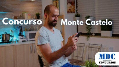 Homem de barba e cabeça raspada, vestindo camiseta cinza e calça azul, sentado em uma cadeira na cozinha, segurando um celular e sorrindo enquanto olha para a tela - Monte Castelo (SP) lança Concurso para Diversas Áreas