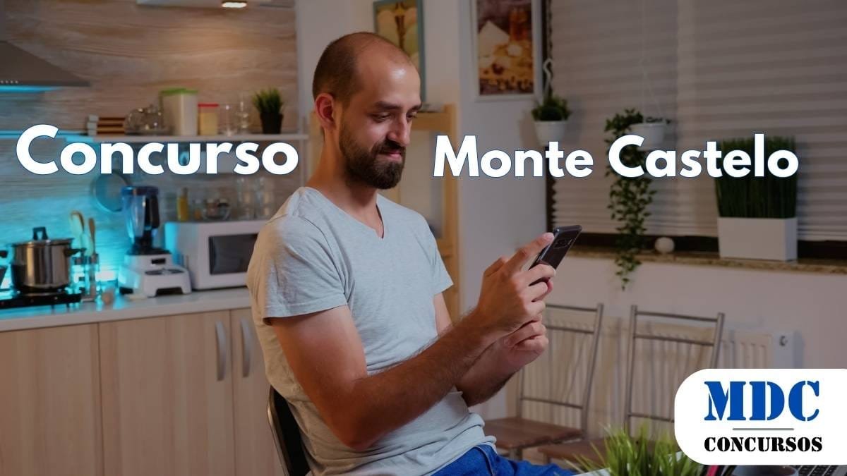 Monte Castelo (SP) lança Concurso para Diversas Áreas 1 Homem de barba e cabeça raspada, vestindo camiseta cinza e calça azul, sentado em uma cadeira na cozinha, segurando um celular e sorrindo enquanto olha para a tela - Monte Castelo (SP) lança Concurso para Diversas Áreas