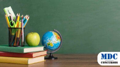 Mesa com materiais escolares diante de um quadro verde. Há um globo terrestre, uma maçã verde, três livros empilhados e um porta-lápis com lápis de cor, canetas, tesoura e régua. O cenário remete ao ambiente escolar, com tons vibrantes e uma composição equilibrada - Morro Cabeça no Tempo (PI) Lança Processo Seletivo