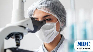 Processos Seletivos 8 Cientista usando touca, máscara e jaleco analisa amostra em um microscópio em um laboratório, com tubos de ensaio desfocados ao fundo - Nova Brasilândia D'Oeste (RO) publica Resultado Preliminar de Processo Seletivo