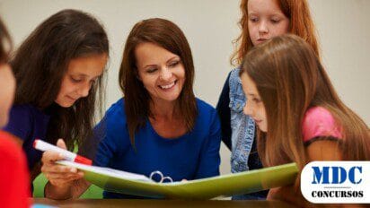Imagem gerada por IA mostrando uma professora sorridente com blusa azul interagindo com quatro alunas em uma sala de aula. Elas estão reunidas em torno de uma mesa, observando um fichário aberto. Há livros, uma calculadora e um marcador na mesa. O ambiente sugere um momento de aprendizado colaborativo e envolvente / PSS em Angicos – RN Abre Vagas na Educação; inscrições até quinta (13)