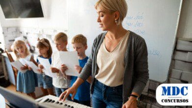Processos Seletivos 4 Professora de música toca teclado enquanto orienta um grupo de crianças que seguram partituras e cantam. A cena acontece em uma sala de aula com quadro branco ao fundo - PSS em Chapecó (SC) abre cadastro de reserva com remuneração de até R$ 4,9 mil