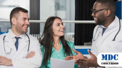 Três profissionais da saúde estão sorrindo e conversando em um ambiente hospitalar. Dois homens usam jalecos brancos e estetoscópios, enquanto a mulher veste um uniforme verde e segura documentos. Eles parecem engajados em uma discussão amigável e profissional, demonstrando um ambiente de trabalho colaborativo / Porto Seguro (BA) Abre 710 Vagas em Processo Seletivo com Salários de até R$ 15 mil