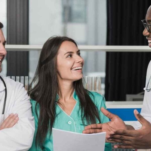 Três profissionais da saúde estão sorrindo e conversando em um ambiente hospitalar. Dois homens usam jalecos brancos e estetoscópios, enquanto a mulher veste um uniforme verde e segura documentos. Eles parecem engajados em uma discussão amigável e profissional, demonstrando um ambiente de trabalho colaborativo / Porto Seguro (BA) Abre 710 Vagas em Processo Seletivo com Salários de até R$ 15 mil
