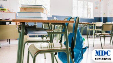 Processos Seletivos 4 Sala de aula vazia com carteiras verdes organizadas. Em primeiro plano, uma mesa com livros empilhados, cadernos e uma mochila azul pendurada na cadeira. Ao fundo, janelas permitem a entrada de luz natural, e as paredes estão decoradas com desenhos infantis e cartazes educativos, criando um ambiente acolhedor e inspirador para o aprendizado / Prefeitura de Amaturá – AM Abre Processo Seletivo com 324 Vagas e Salários de até R$ 4,9 mil