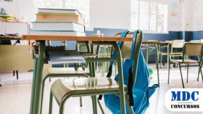 Sala de aula vazia com carteiras verdes organizadas. Em primeiro plano, uma mesa com livros empilhados, cadernos e uma mochila azul pendurada na cadeira. Ao fundo, janelas permitem a entrada de luz natural, e as paredes estão decoradas com desenhos infantis e cartazes educativos, criando um ambiente acolhedor e inspirador para o aprendizado / Prefeitura de Amaturá – AM Abre Processo Seletivo com 324 Vagas e Salários de até R$ 4,9 mil