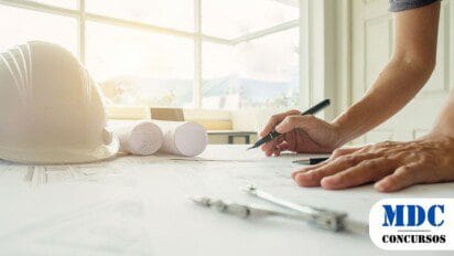 Pessoa trabalha em projeto de arquitetura ou engenharia sobre a mesa, com capacete branco, pranchetas, caneta e compassos. Ambiente iluminado por luz natural / Prefeitura de Catas Altas – MG abre Processo Seletivo com Salários de até R$ 9,6 mil