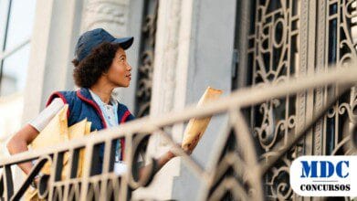 Processos Seletivos 8 Mulher carteiro com uniforme azul e boné entrega envelopes em um prédio com portão ornamental de ferro. Ela segura correspondências e olha para a porta com expressão atenta. A cena ocorre em um ambiente urbano, com arquitetura clássica ao fundo / Prefeitura de Crato – CE abre Processo Seletivo para Cadastro de Reserva