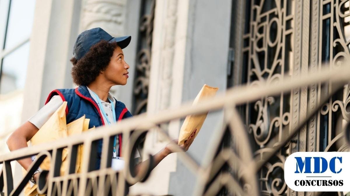 Prefeitura de Crato – CE abre Processo Seletivo para Cadastro de Reserva 1 Mulher carteiro com uniforme azul e boné entrega envelopes em um prédio com portão ornamental de ferro. Ela segura correspondências e olha para a porta com expressão atenta. A cena ocorre em um ambiente urbano, com arquitetura clássica ao fundo / Prefeitura de Crato – CE abre Processo Seletivo para Cadastro de Reserva