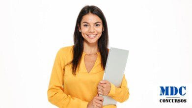 Mulher jovem de cabelos castanhos e longos, vestindo blusa amarela com detalhe dourado na gola, segurando um laptop prateado e sorrindo. Fundo branco, expressão amigável e postura confiante - Prefeitura de Patos (PB) Lança Processo Seletivo com 215 Vagas