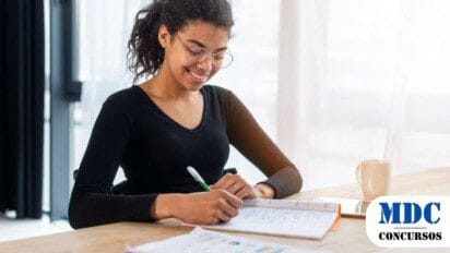 Mulher jovem de óculos e cabelos cacheados presos, vestindo uma blusa preta, sorrindo enquanto escreve em um caderno. Ela está sentada à mesa com papéis, um celular e uma xícara ao lado. O ambiente é iluminado e moderno, com cortinas brancas ao fundo - Prefeitura de Piracuruca (PI) lança Processo Seletivo com 40 Vagas na Educação