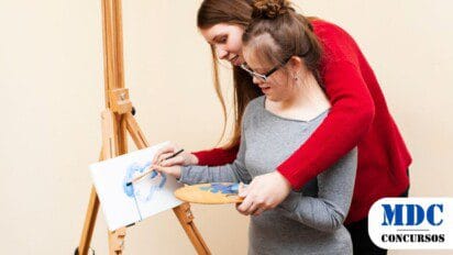 Duas jovens pintam juntas em um cavalete. A mulher de vermelho ajuda a outra, segurando suas mãos enquanto ela usa um pincel para aplicar tinta azul na tela. A jovem de cinza segura uma paleta com várias cores. Ambas parecem felizes e concentradas. O fundo é simples e claro, destacando o momento artístico e de aprendizado / Prefeitura de Rio Verde – GO abre 2 Processos Seletivos com 165 Vagas
