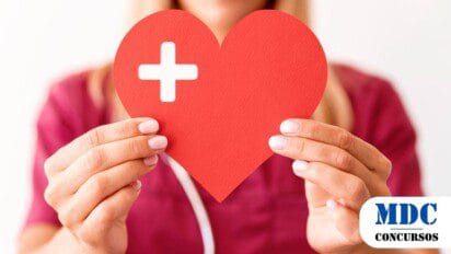Mulher com uniforme de profissional de saúde segura um coração vermelho recortado de papel com um símbolo de cruz branca no canto, representando cuidado e saúde - Prefeitura de Sarzedo (MG) Oferece 58 Vagas em 2 Processos Seletivos