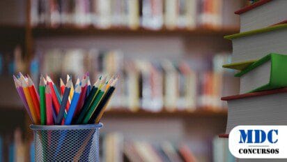 Porta-lápis metálico com lápis de cor variados em primeiro plano e uma pilha de livros com marcadores coloridos ao lado, contra um fundo desfocado de estantes de livros - Prefeitura de Teixeira (PB) Abre 200 Vagas para Educador Social